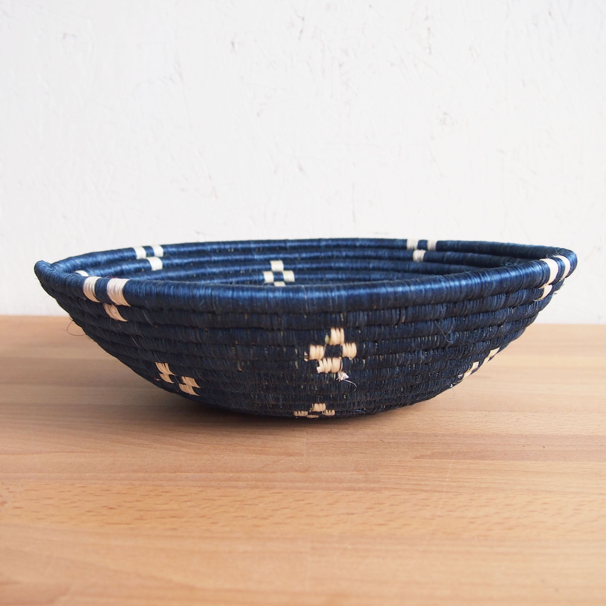 The Kabaya Large Bowl by Amsha - Faire, a round, shallow basket woven from dark blue and natural fibers with light geometric accents, rests on a wooden surface against a white textured wall.