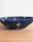 The Kabaya Large Bowl by Amsha - Faire, a round, shallow basket woven from dark blue and natural fibers with light geometric accents, rests on a wooden surface against a white textured wall.