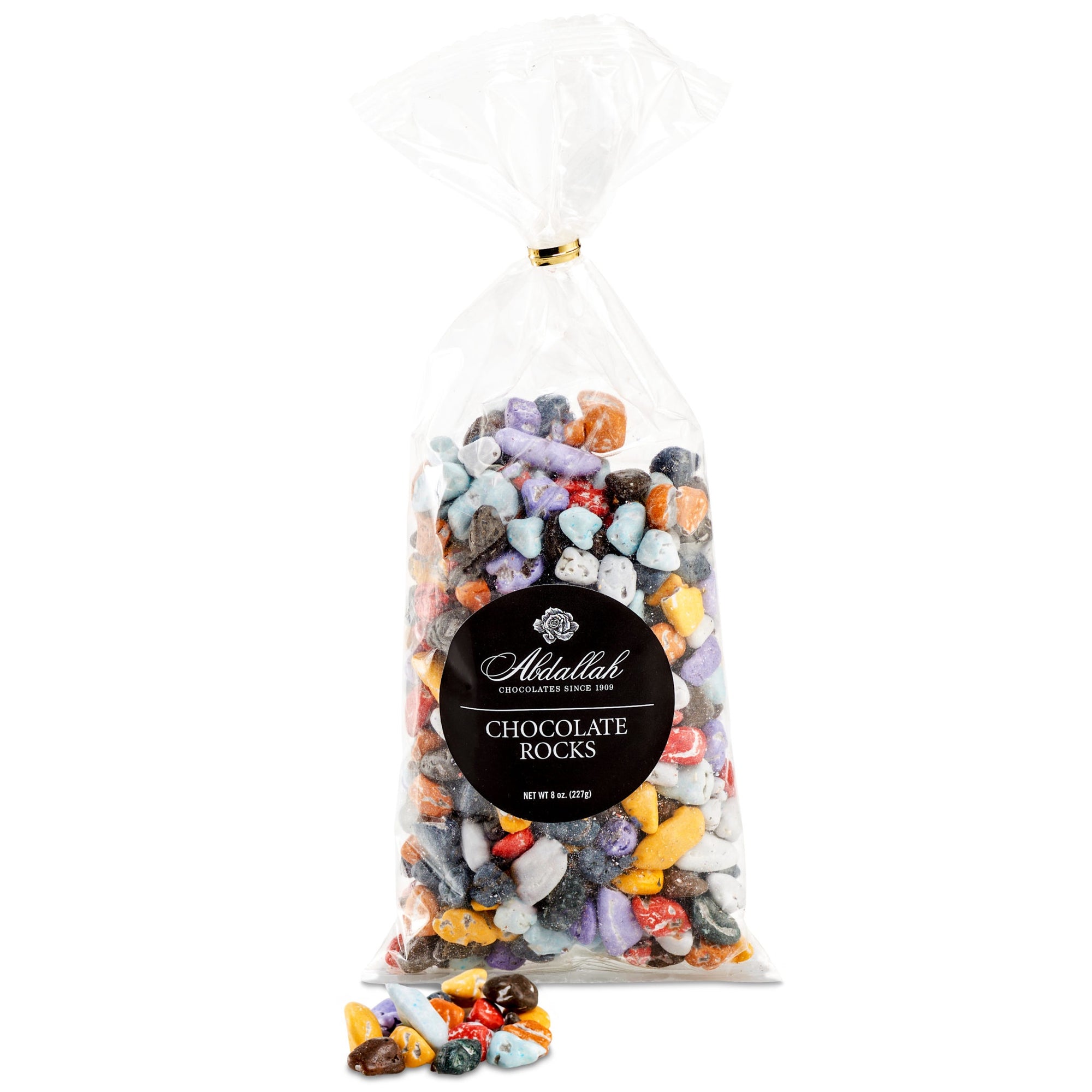 A clear plastic bag filled with vibrant Abdallah Choco Rocks, candy-coated milk chocolates, stands upright. The label on the bag reads "Choco Rocks," with a few colorful chocolate rocks scattered at the base like river stones.