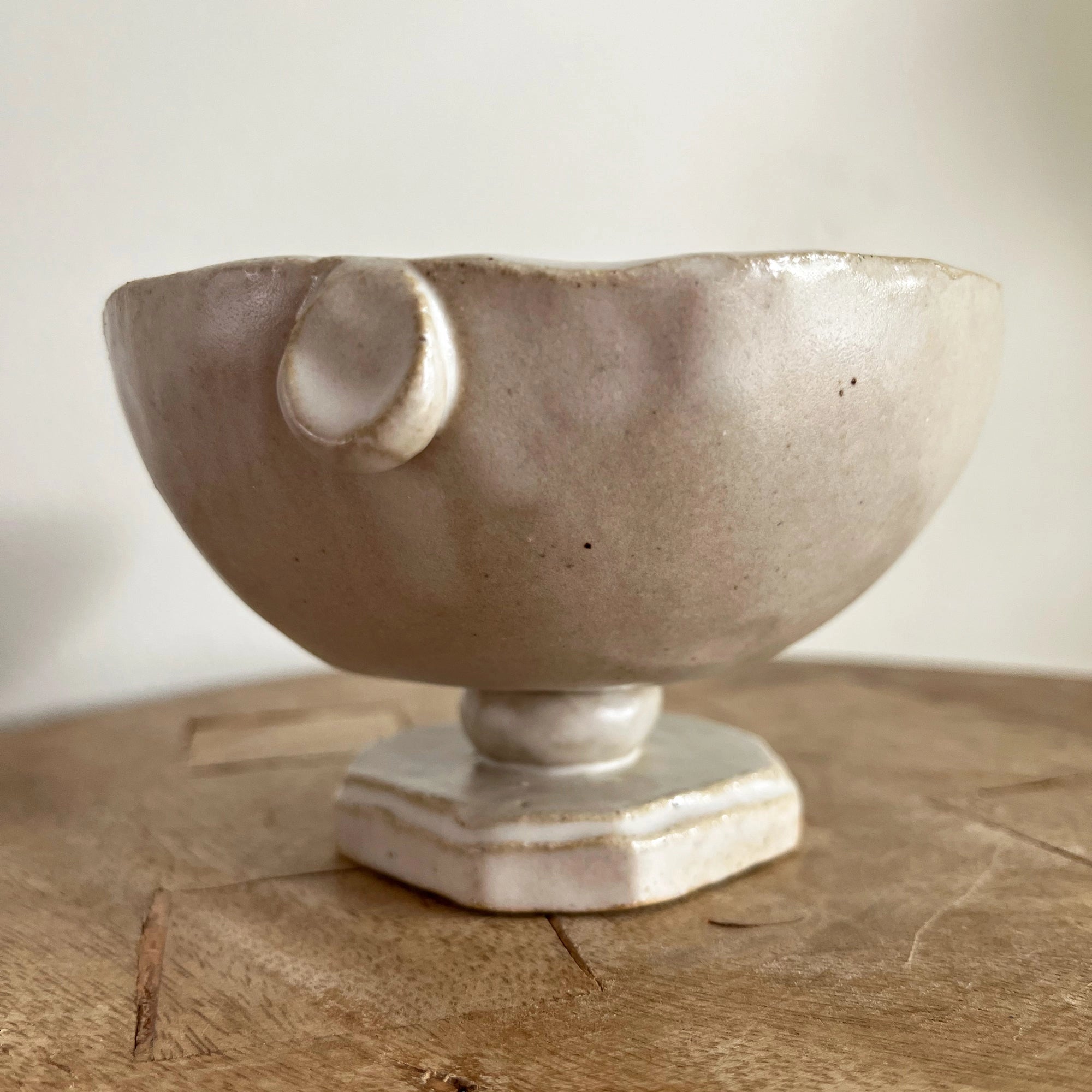 The Faire Tiny Trophy Bowl, a handmade glazed stoneware trinket dish with an off-white finish, irregular rim, small handle detail, and hexagonal pedestal base, rests on a wooden surface against a plain background.