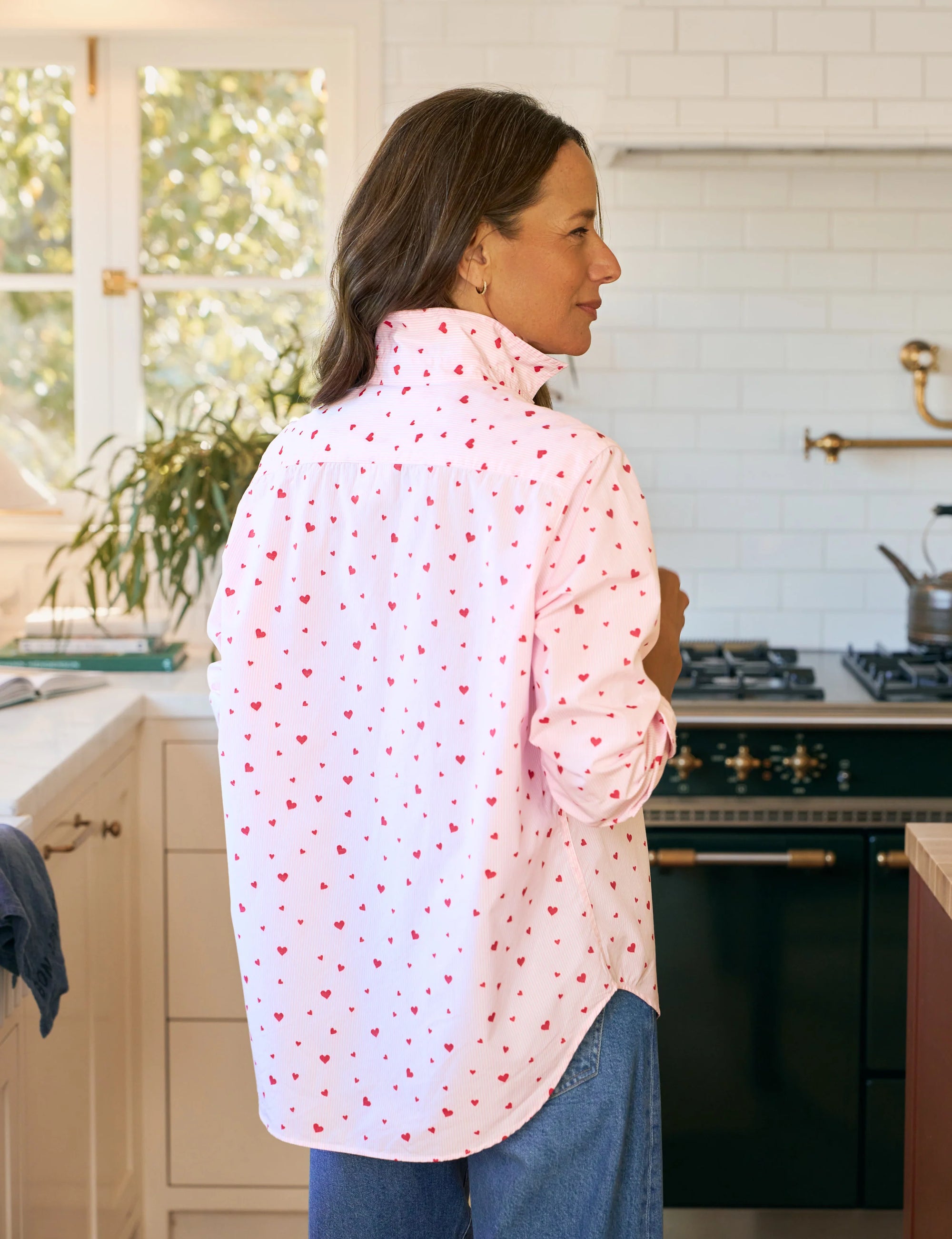 A woman stands in a sunlit kitchen wearing the Frank & Eileen Eileen Tops 2026, a relaxed-fit shirt with small red hearts, paired with blue jeans. Sunlight highlights a green plant on the countertop as she faces away.