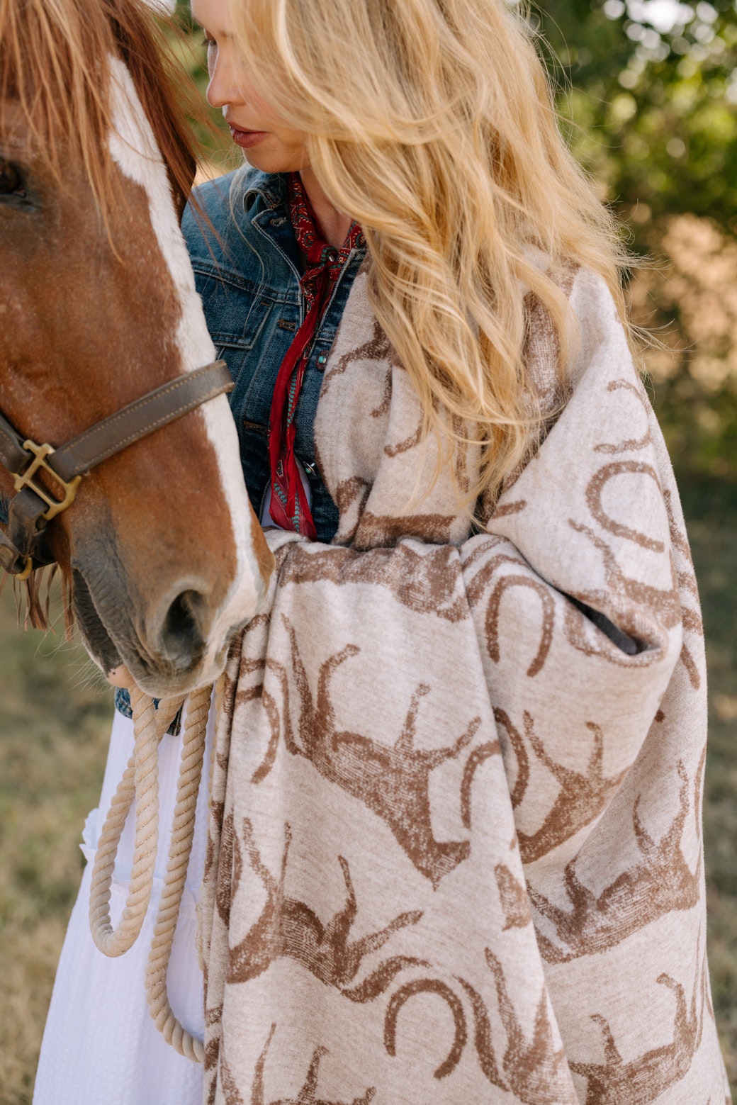 A woman with long blonde hair stands outdoors among trees, wrapped in the Faire Hill Country Horses Lightweight Blanket featuring horse and horseshoe patterns. She wears a denim jacket and red scarf while holding a brown horse closely.
