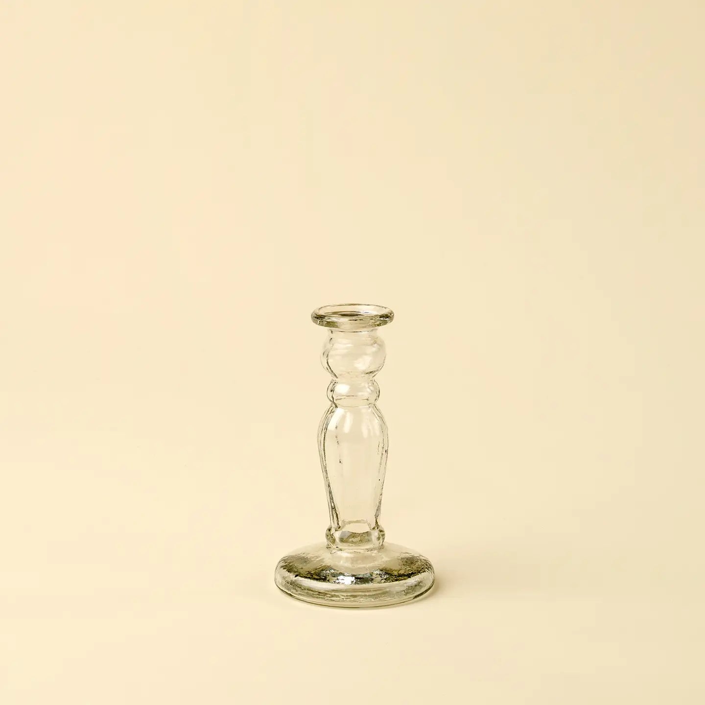 The Vero Glass Candlestick S by Indaba features a slender, rounded stem and is crafted from handblown recycled glass, offering a sustainable and timeless candle holder. Displayed on a plain, cream-colored background.