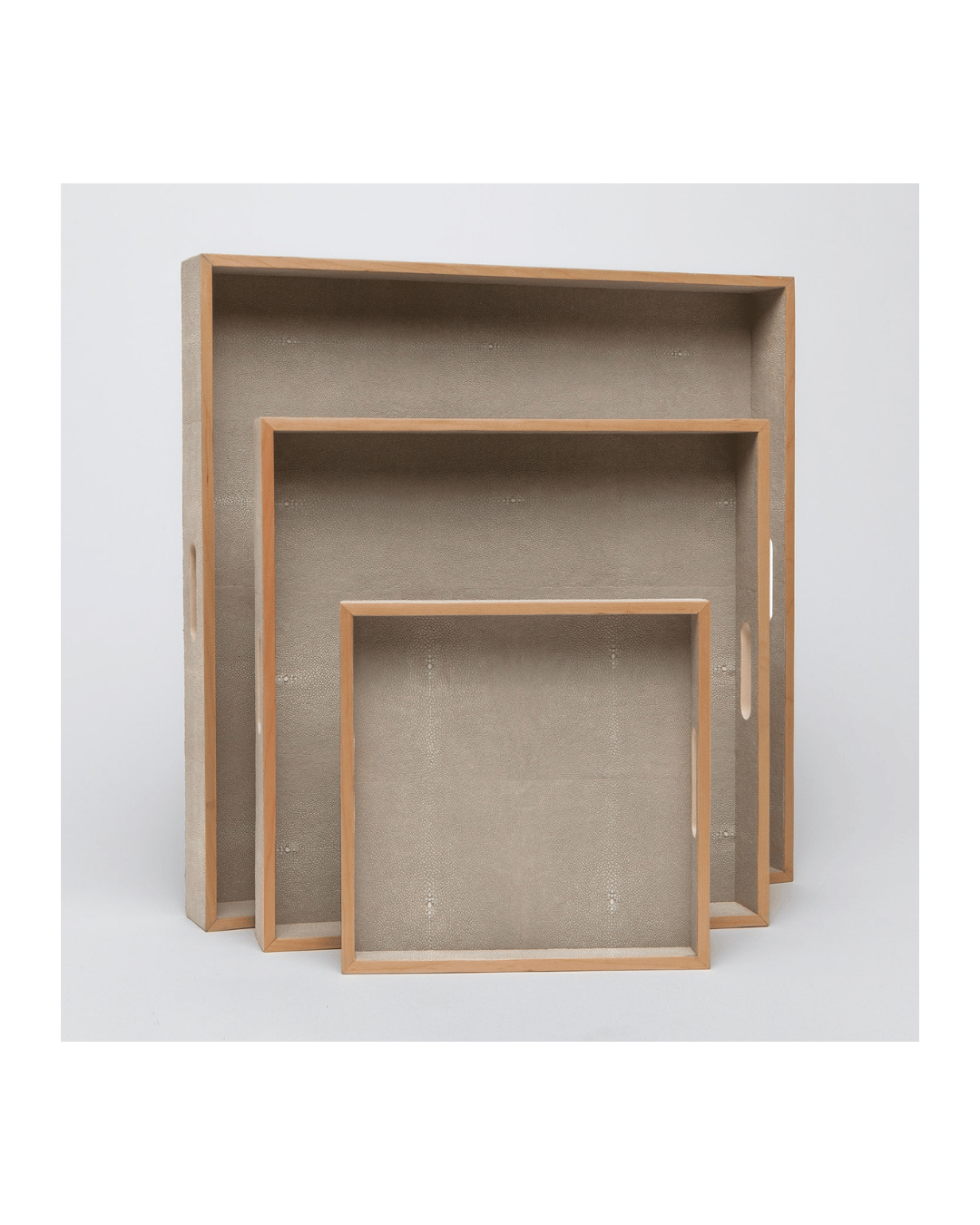 The Amina Tray by Made Goods, showcasing three nested trays of varying sizes with elegant wood trim, is displayed against a plain white background. The trays are positioned upright with the largest at the back and the smallest in front, highlighting their rectangular shapes and simple, natural wood finish.