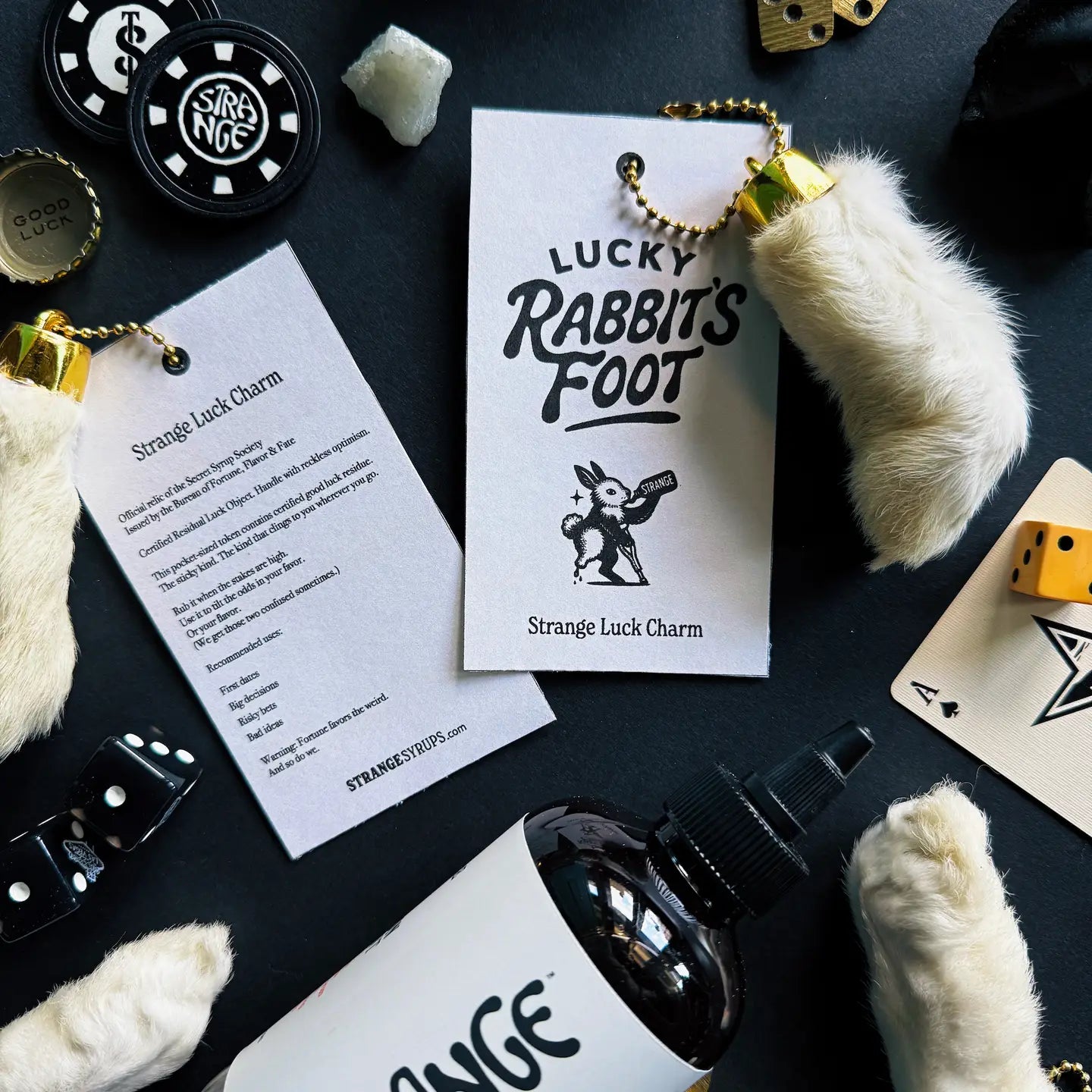 A flat lay of Faire’s Lucky Rabbit's Foot keychains tagged “Strange Luck Charm,” surrounded by dice, poker chips, cards, and a “STRANGE” bottle—the perfect good luck charm for gamblers.