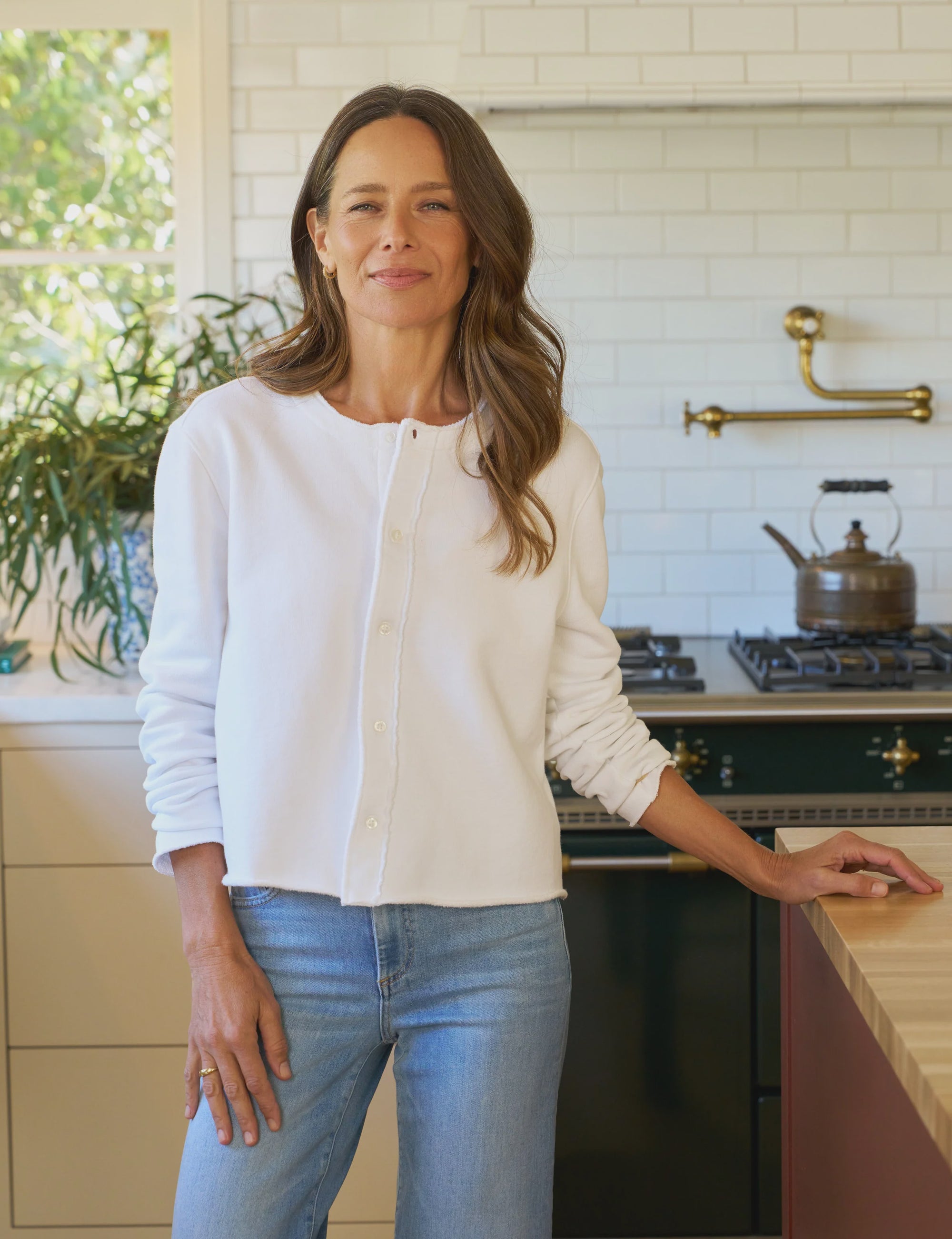 A woman with long brown hair smiles in a bright kitchen, wearing the Frank & Eileen Jackie Sweatshirt Cardigan over a white shirt and blue jeans; white subway tile, stove, kettle, and plants are visible in the background.