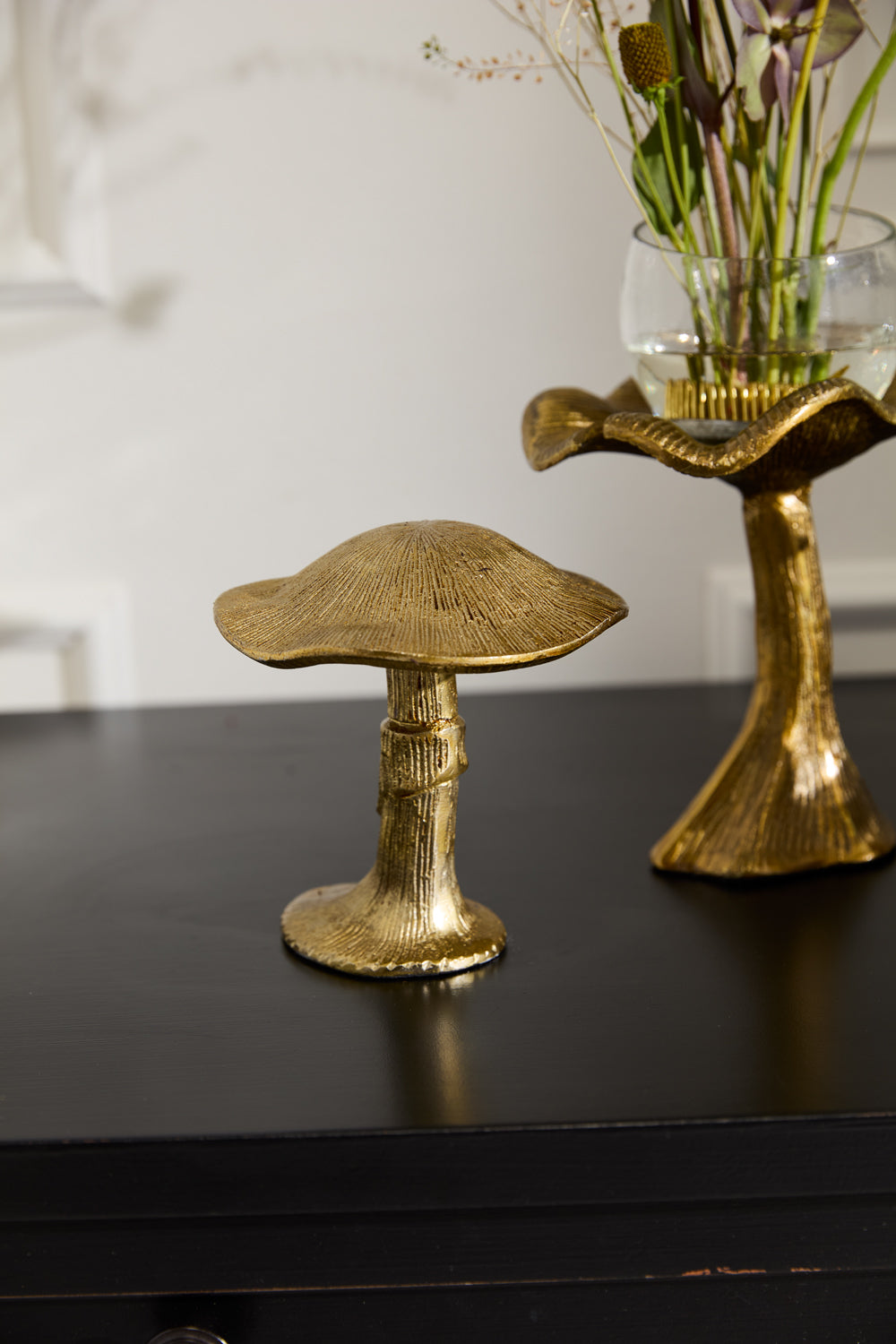 The Lichen Figurine by Accent Decor, a gold metal mushroom sculpture, sits on a black surface beside a matching vase with flowers. This nature-inspired decor stands out against a light-colored background with subtle wall details.