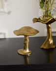 The Lichen Figurine by Accent Decor, a gold metal mushroom sculpture, sits on a black surface beside a matching vase with flowers. This nature-inspired decor stands out against a light-colored background with subtle wall details.