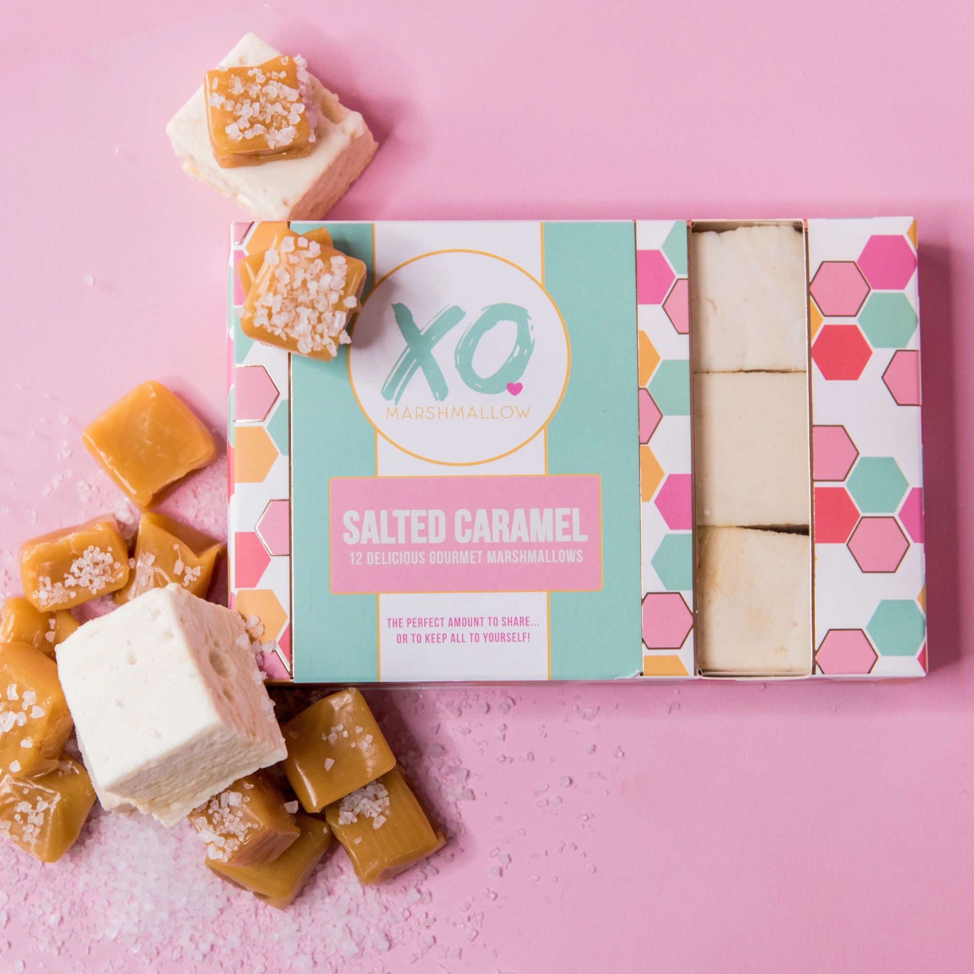 A box of Faire Salted Caramel Marshmallows rests on a pink surface, with small batch marshmallows and caramel pieces sprinkled with sea salt arranged beside it.