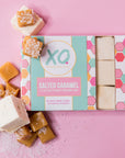 A box of Faire Salted Caramel Marshmallows rests on a pink surface, with small batch marshmallows and caramel pieces sprinkled with sea salt arranged beside it.