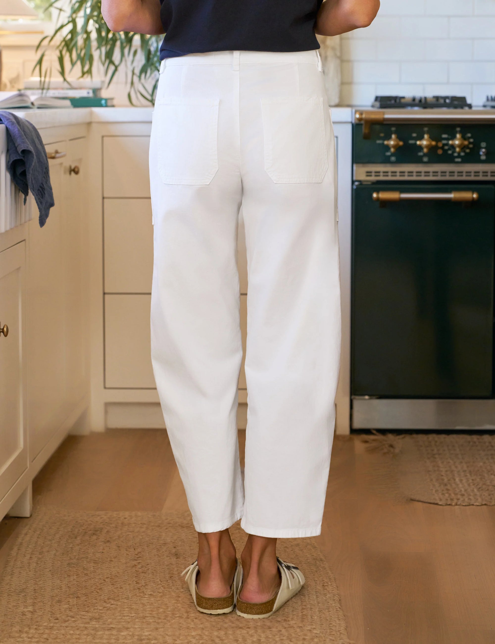 A person stands in a kitchen wearing the Frank & Eileen Everyday Barrel Pant in white and beige slip-on shoes. The kitchen has a green stove, white cabinets, and wooden floors.