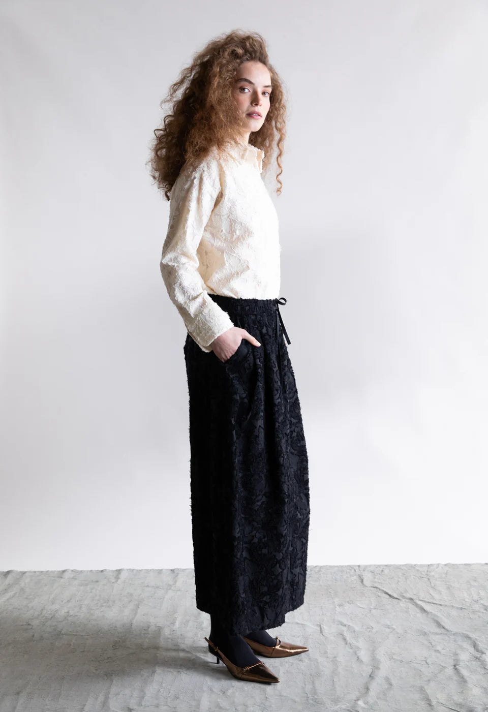A woman with curly hair stands sideways against a plain background, wearing the Psophia Embroidered Shirt tucked into a long black patterned skirt and pointed brown heels, her hands in her skirt pockets.