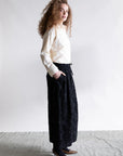 A woman with curly hair stands sideways against a plain background, wearing the Psophia Embroidered Shirt tucked into a long black patterned skirt and pointed brown heels, her hands in her skirt pockets.