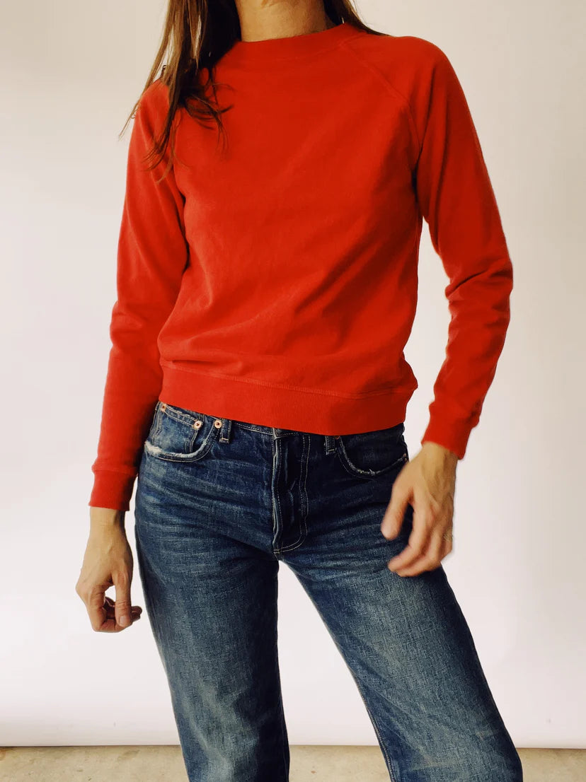 A person stands casually, wearing &quot;The Sweatshirt&quot; by Hey Gang, paired with blue jeans. Their right hand is slightly raised against a plain, off-white wall. The image captures a relaxed and comfortable style characteristic of classic fashion moments.