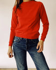 A person stands casually, wearing "The Sweatshirt" by Hey Gang, paired with blue jeans. Their right hand is slightly raised against a plain, off-white wall. The image captures a relaxed and comfortable style characteristic of classic fashion moments.