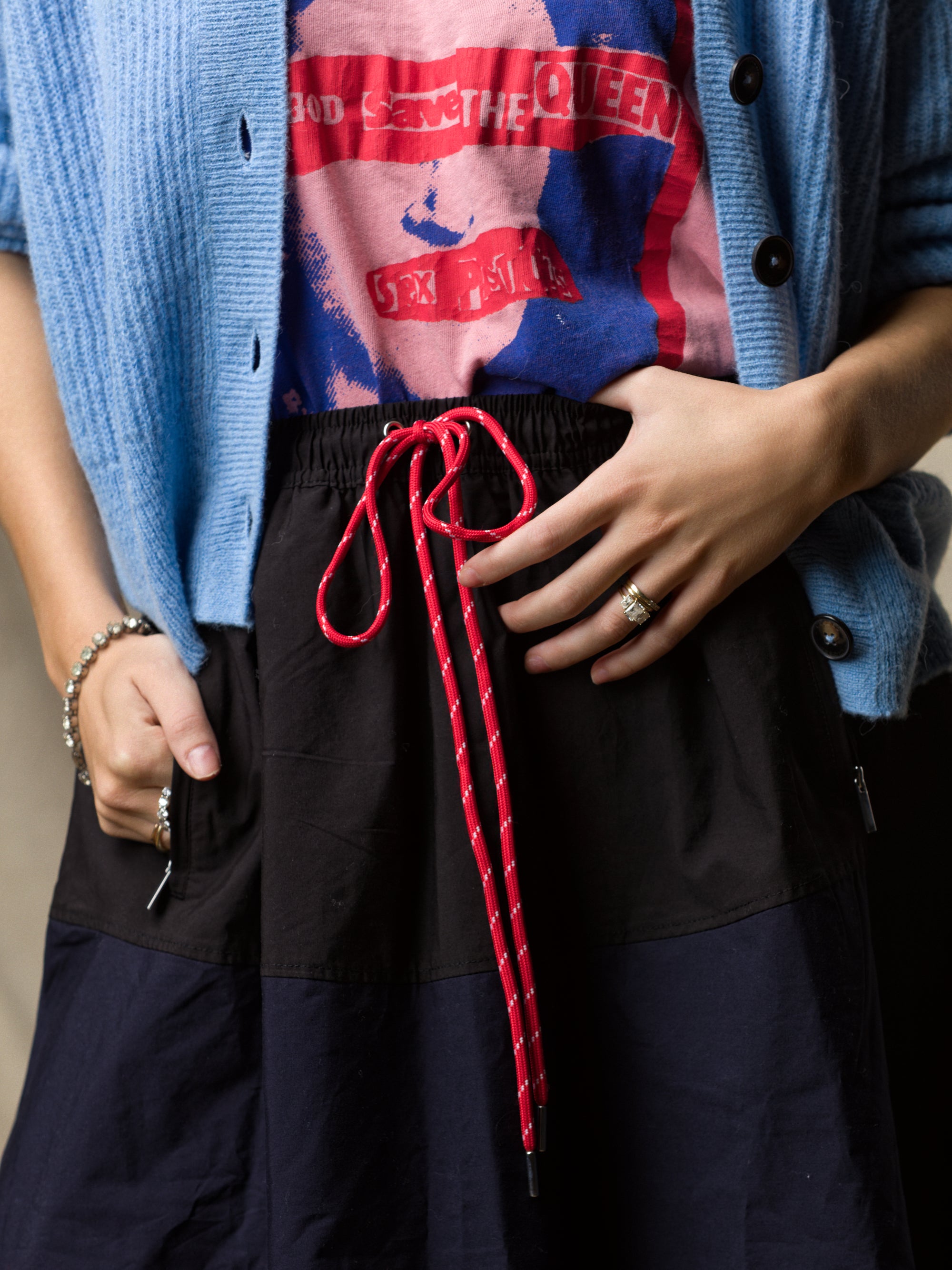 Wearing a blue cardigan, a red and blue graphic tee, and the Ping Pong Surf Club Le Vintage Sport Skirt with red rope belt, this look—completed by ring and bracelet—captures signature Ping Pong Surf Club style.