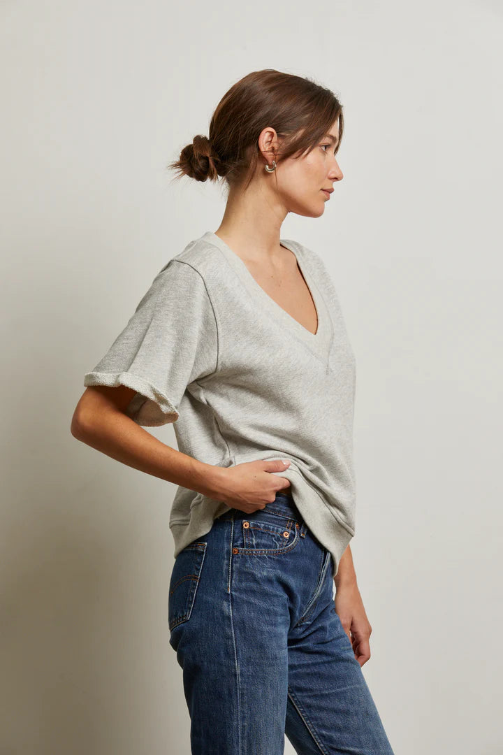A woman with brown hair in a low bun, wearing the Perfectwhitetee French Terry SS Crew in light gray and blue jeans, stands in profile against a plain off-white background.