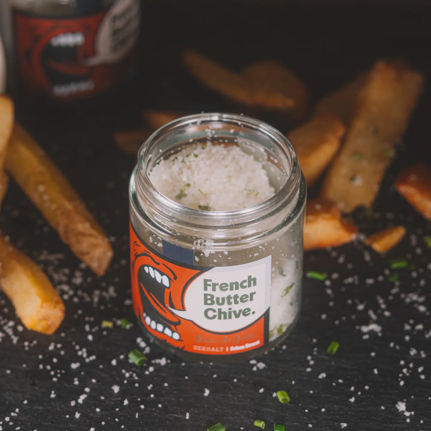 A jar labeled "Fleur de Sel Sea Salt" by Faire is surrounded by French fries on a dark surface, with scattered salt and chopped chives. The jar features a graphic illustration of an open mouth, with coarse salt sprinkled on top.