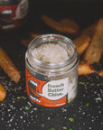 A jar labeled "Fleur de Sel Sea Salt" by Faire is surrounded by French fries on a dark surface, with scattered salt and chopped chives. The jar features a graphic illustration of an open mouth, with coarse salt sprinkled on top.