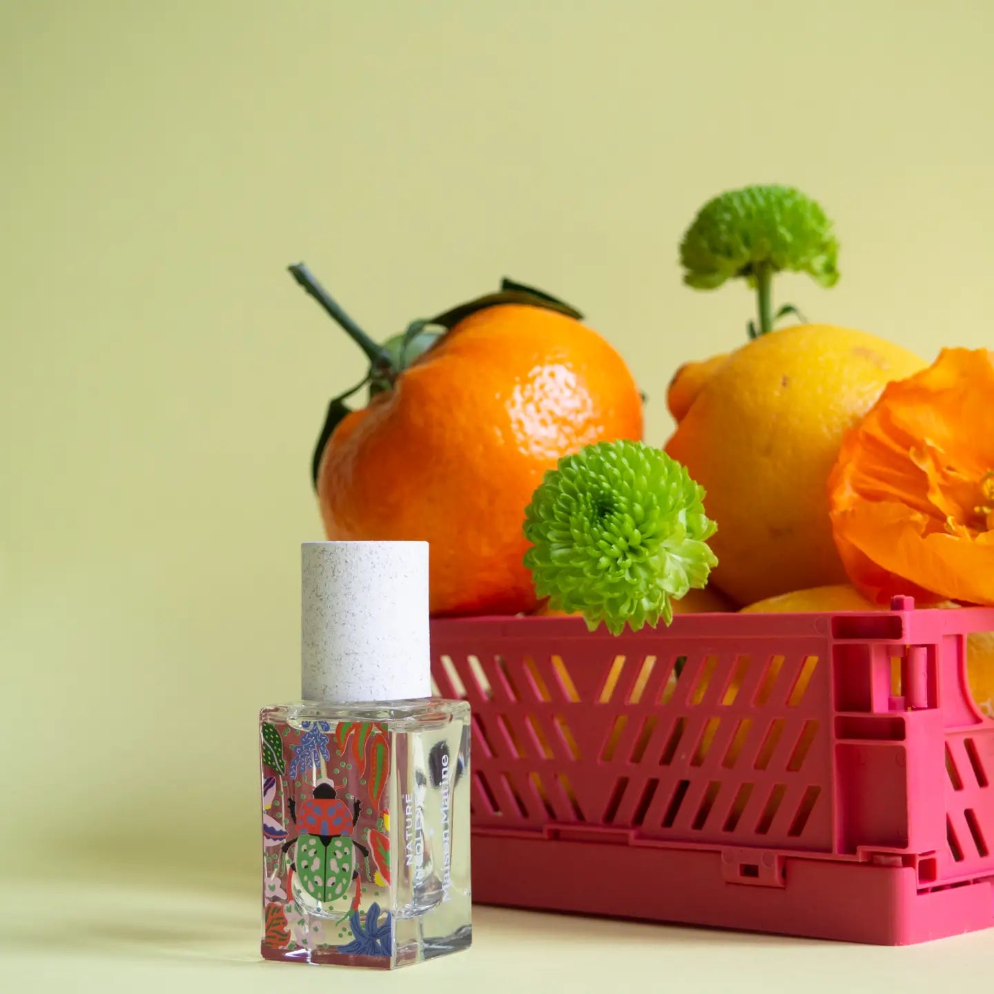 The Maison Matine Perfume by Faire features a colorful bottle with a white cap beside a pink crate filled with citrus fruits, vibrant flowers, and bergamot accents, all set against a pale yellow background.