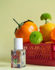 The Maison Matine Perfume by Faire features a colorful bottle with a white cap beside a pink crate filled with citrus fruits, vibrant flowers, and bergamot accents, all set against a pale yellow background.
