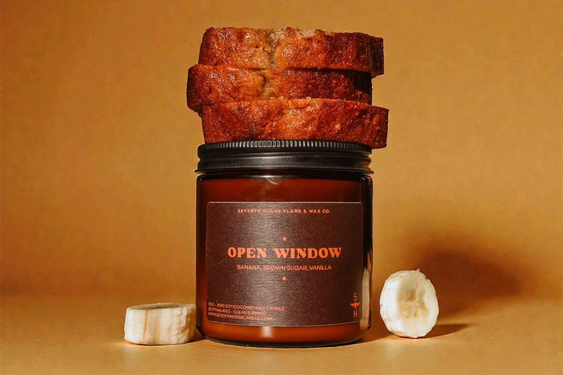 A Faire Banana Nut Bread Candle in a brown glass jar labeled "Open Window" sits on a tan surface with banana bread slices and banana pieces beside it, set against a warm brown background.