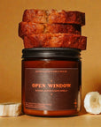 A Faire Banana Nut Bread Candle in a brown glass jar labeled "Open Window" sits on a tan surface with banana bread slices and banana pieces beside it, set against a warm brown background.