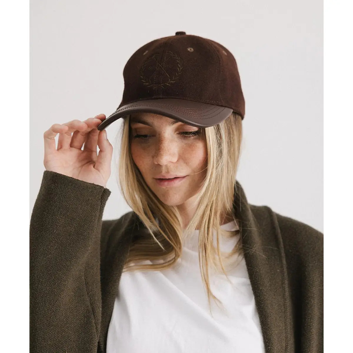 A person with long blonde hair wears the Faire Aven Leather Ball Cap, styled with an olive green jacket and white shirt, looking downward while touching the cap’s brim against a plain light background.