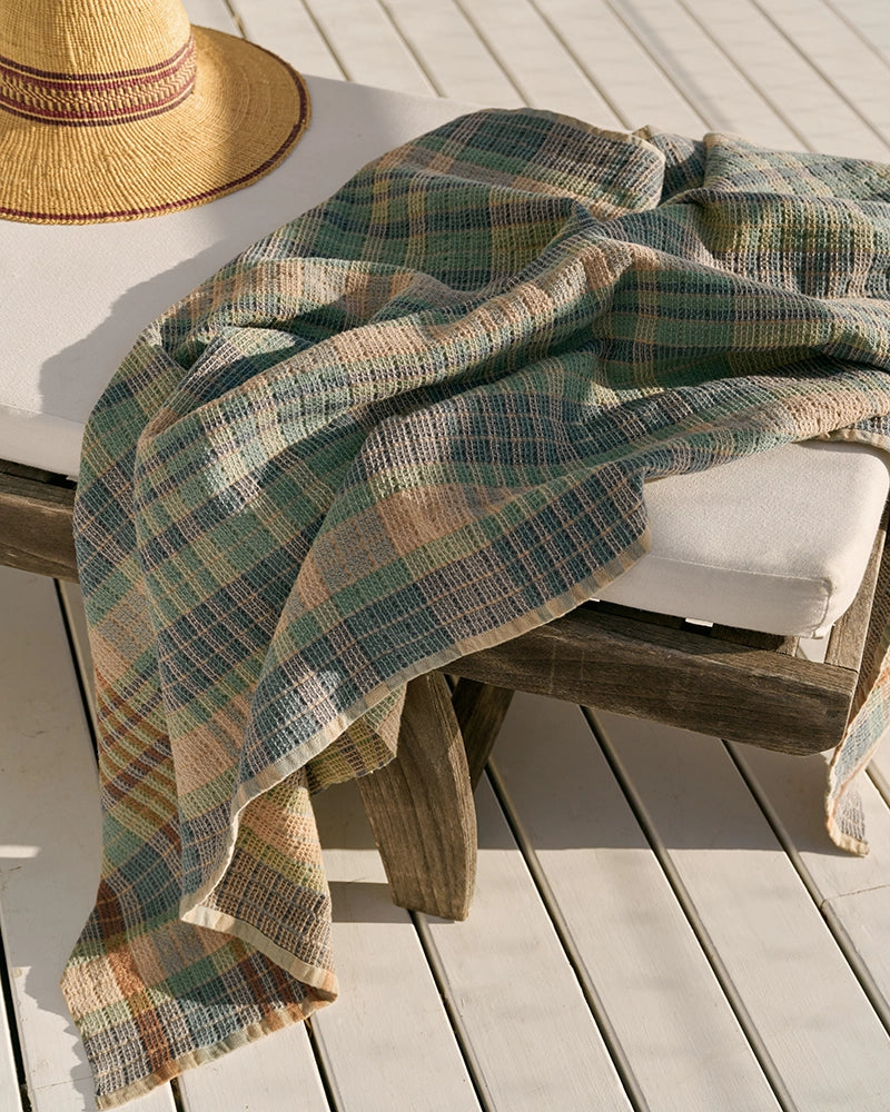 A plaid blanket covers a cushioned wooden lounge chair beside the Faire Wayfarer Towel and a wide-brimmed straw hat, all arranged on a sunlit wooden deck.
