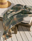 A plaid blanket covers a cushioned wooden lounge chair beside the Faire Wayfarer Towel and a wide-brimmed straw hat, all arranged on a sunlit wooden deck.