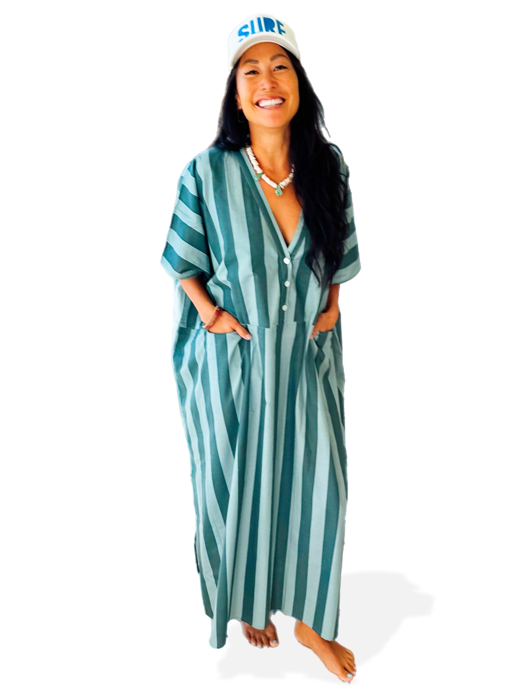 A smiling woman with long dark hair stands barefoot in a blue and teal Faire Suft Stripe Kaftan, a lightweight cotton kaftan with coastal vibes. She wears a white "SURF" cap and necklace against a plain white background.