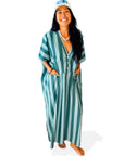 A smiling woman with long dark hair stands barefoot in a blue and teal Faire Suft Stripe Kaftan, a lightweight cotton kaftan with coastal vibes. She wears a white "SURF" cap and necklace against a plain white background.