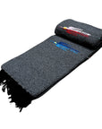 The Charcoal Thunderbird Baja Blanket by Faire is a handmade gray woven blanket with black fringe and two embroidered horses—one red, one blue—near the top edge, inspired by classic Mexican Thunderbird blanket designs.