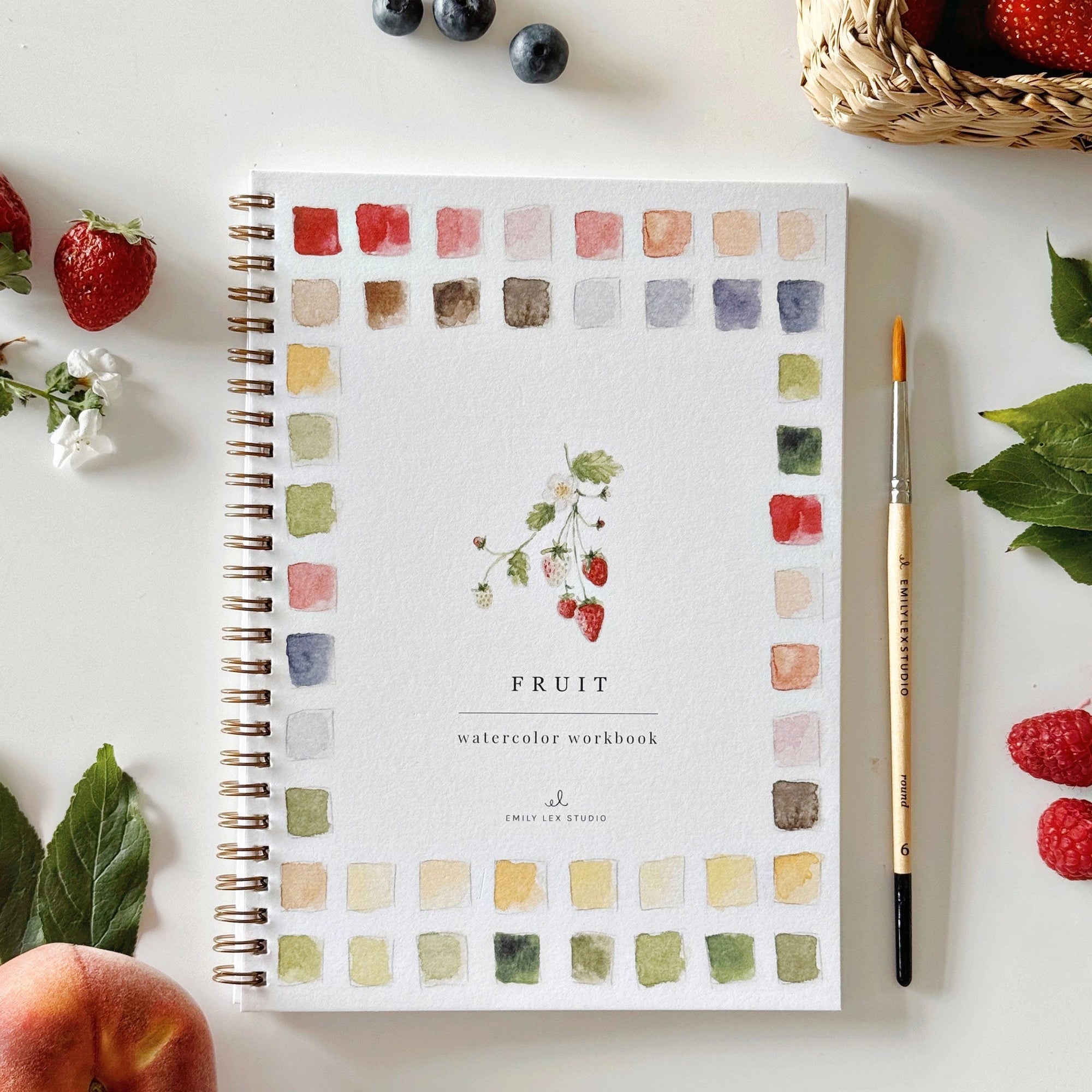The Faire Watercolor Workbook is a spiral-bound coloring book featuring fruit illustrations, ideal for beginner watercolor artists, shown with a paintbrush, leaves, berries, and a woven basket on a white surface.