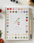 The Faire Watercolor Workbook is a spiral-bound coloring book featuring fruit illustrations, ideal for beginner watercolor artists, shown with a paintbrush, leaves, berries, and a woven basket on a white surface.