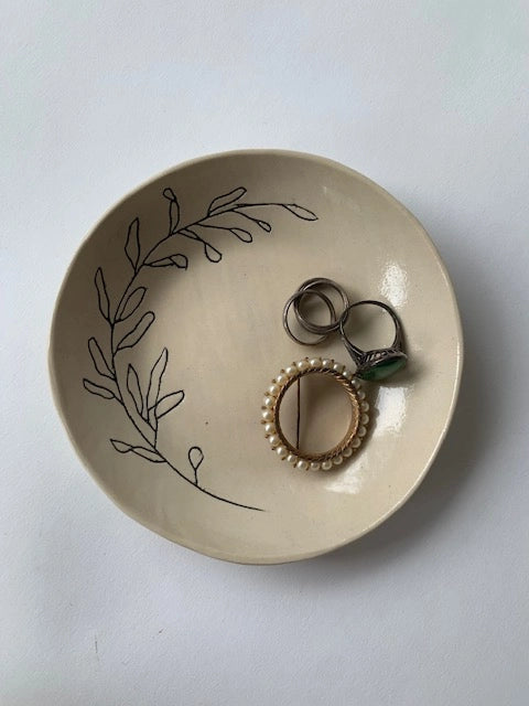 The Faire Ring Bowl, a cream hand-etched tray with a black leafy design, holds a pearl brooch and rings, serving as a chic jewelry catch-all against a white background.
