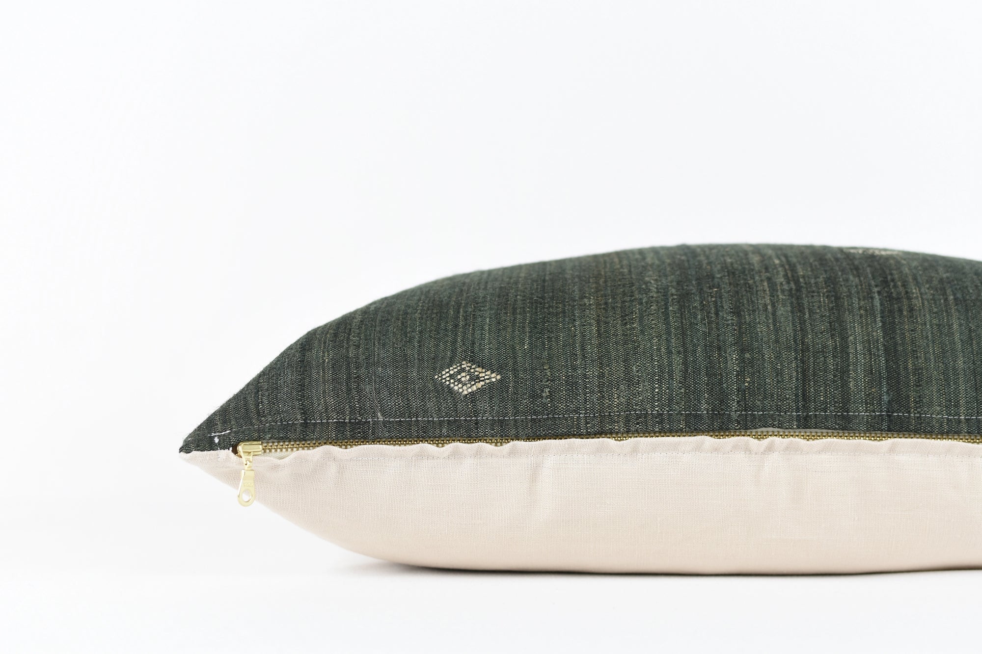 The Dahlia Black 22x22 Pillow by Faire features handwoven dark green silk textured fabric on top, a small stitched detail, beige bottom, and gold zipper, inspired by Gujarat craftsmanship. Shown here against a plain white background.
