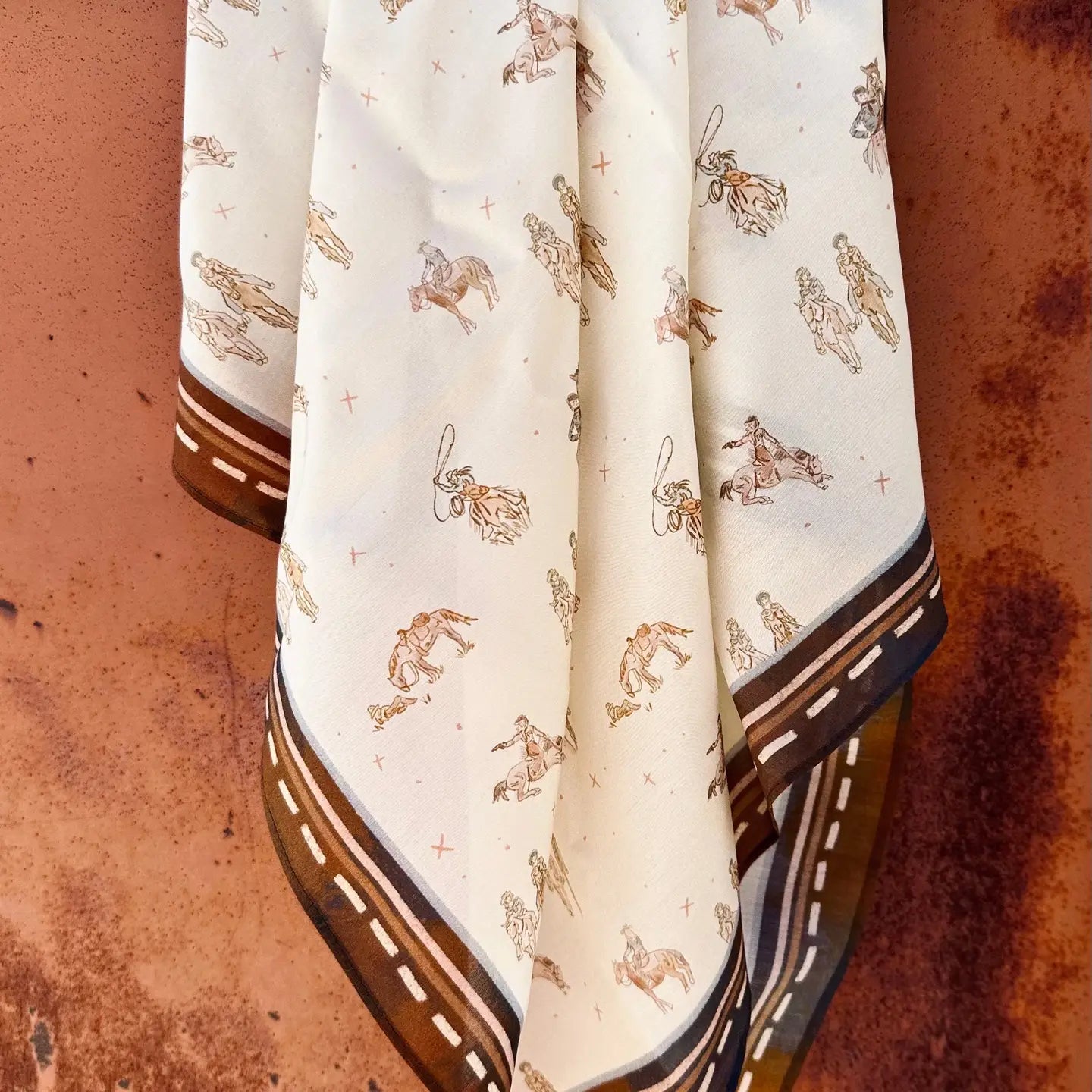 The Faire Wild Rag Silk Scarf is cream-colored with brown and tan cowboy and horse designs, a dark brown border with dashed white lines, and is styled here against a rustic metal backdrop.
