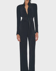 Dressed in the NK Shirt Straight Leg Jumpsuit by Norma Kamali, a person poses confidently against a plain white background. The black jumpsuit features long sleeves, 4-way stretch fabric, shoulder pads, and is elegantly paired with a belt and sleek black shoes.