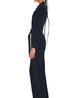 A person with braided hair stands in profile against a white background, wearing the NK Shirt Straight Leg Jumpsuit by Norma Kamali, complete with shoulder pads and a belt, complemented by sleek black shoes.