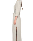 A woman with a long braid stands in profile, barefoot, modeling the Norma Kamali Bomber Long Dress—a vintage-inspired, light gray ankle-length piece with crew neckline, long sleeves, fabric belt, and high side slit—against a plain white background.