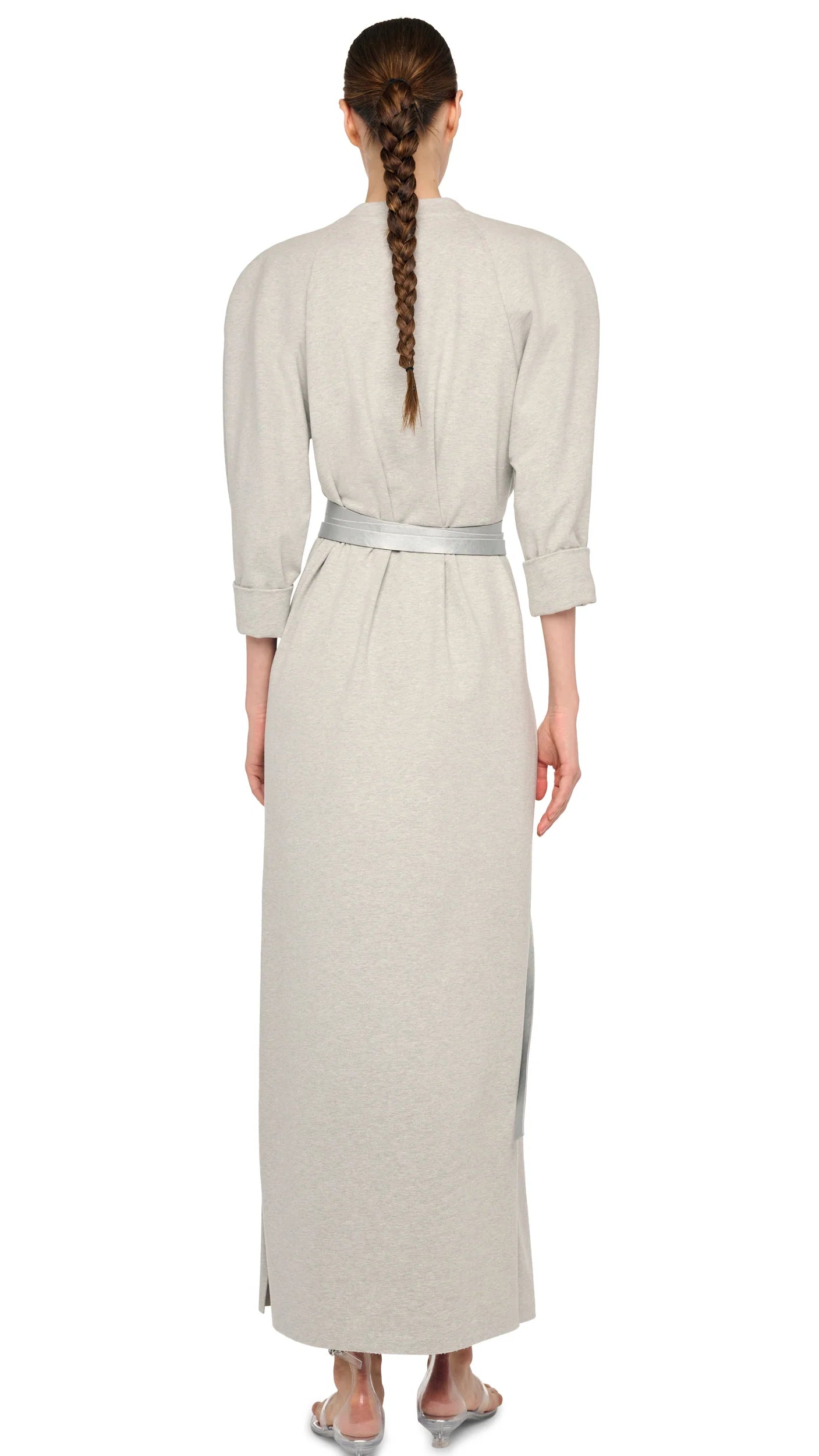 A woman with a long braid is seen from the back wearing Norma Kamali’s Bomber Long Dress, a vintage-inspired, light grey, long-sleeved, ankle-length piece with a crew neckline and fabric belt, paired with flat sandals.