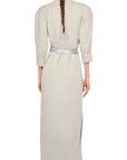 A woman with a long braid is seen from the back wearing Norma Kamali’s Bomber Long Dress, a vintage-inspired, light grey, long-sleeved, ankle-length piece with a crew neckline and fabric belt, paired with flat sandals.