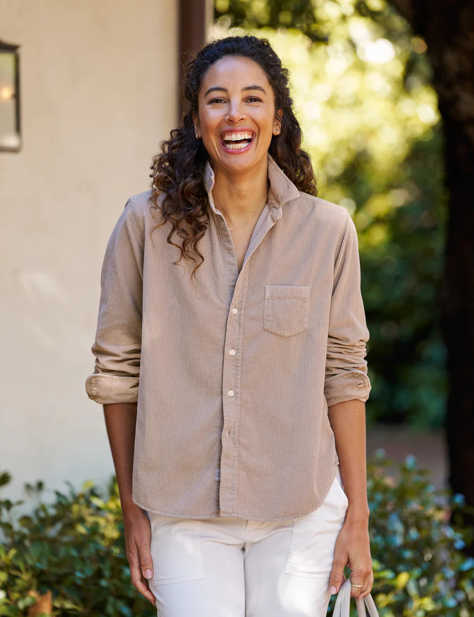 A woman with curly hair smiles outdoors in a Frank & Eileen EILEEN Relaxed Button-Up Shirt and white pants, standing before greenery and holding a light-colored bag.