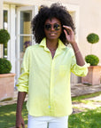 A person with curly hair stands outdoors, adjusting their sunglasses while smiling. They wear a bright yellow Eileen S25, an Italian cotton button-up shirt by Frank & Eileen. The relaxed fit complements the scene of trees and a building with glass doors in the background.