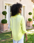 A person with curly hair, wearing sunglasses and a relaxed-fit, light yellow Eileen S25 shirt by Frank & Eileen, stands outside near potted spherical shrubs and a glass door. They are slightly turned, facing the garden, exuding an effortless vibe.