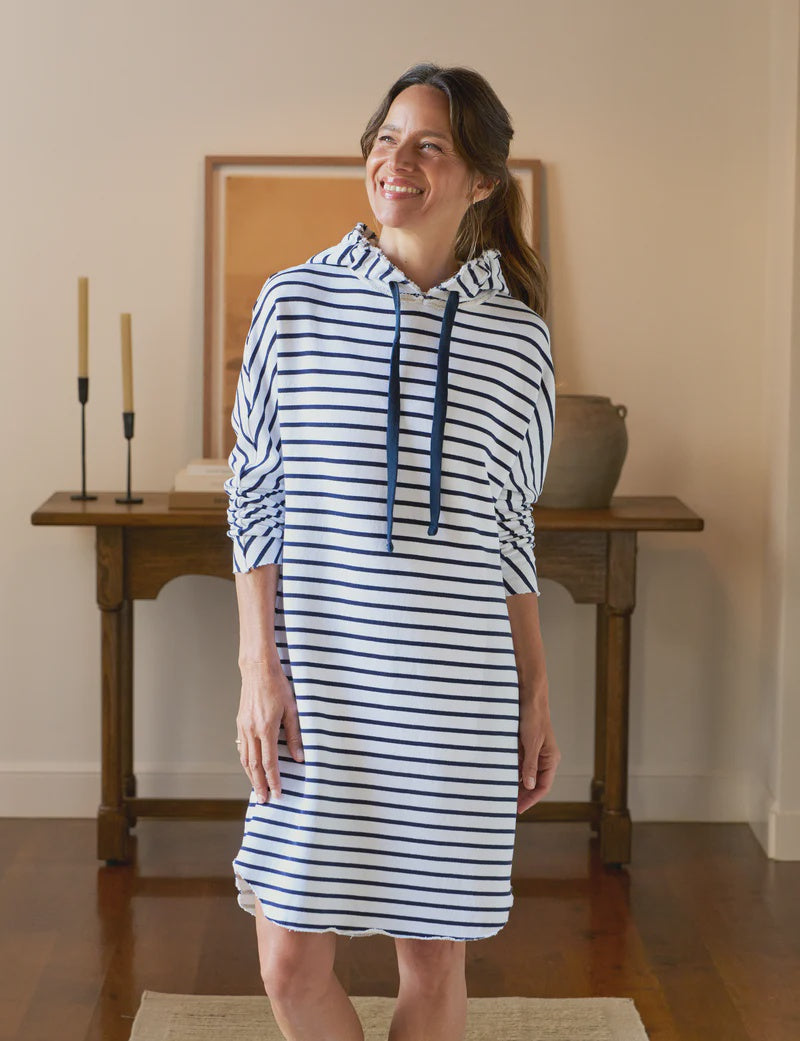 A smiling woman stands indoors wearing the Mini James Hoodie Cape Dress by Frank & Eileen—a blue-and-white striped, drawstring mini dress. She is in a warmly lit room with wooden floors, a table, candles, a vase, and framed artwork.