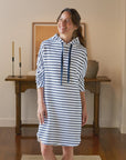 A smiling woman stands indoors wearing the Mini James Hoodie Cape Dress by Frank & Eileen—a blue-and-white striped, drawstring mini dress. She is in a warmly lit room with wooden floors, a table, candles, a vase, and framed artwork.