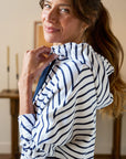 A woman with long brown hair in a ponytail smiles gently indoors, holding the collar of her Frank & Eileen Mini James Hoodie Cape Dress—a white mini pullover dress with navy blue stripes—against a softly blurred backdrop.