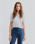 A woman with long wavy brown hair wears The Farley short-sleeve polo sweater by Kule, a light gray cotton-silk blend, paired with high-waisted blue pants, standing against a plain light background.